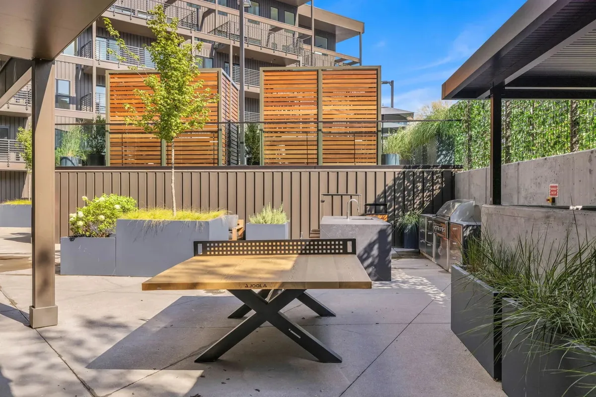 An outdoor grilling and dining area—featuring a large wooden table, grilling station, and privacy-enhancing wooden screens—invites groups to share meals and laughter al fresco. Planters with greenery add a touch of serenity, while the communal setting suggests festive gatherings and barbecue nights.