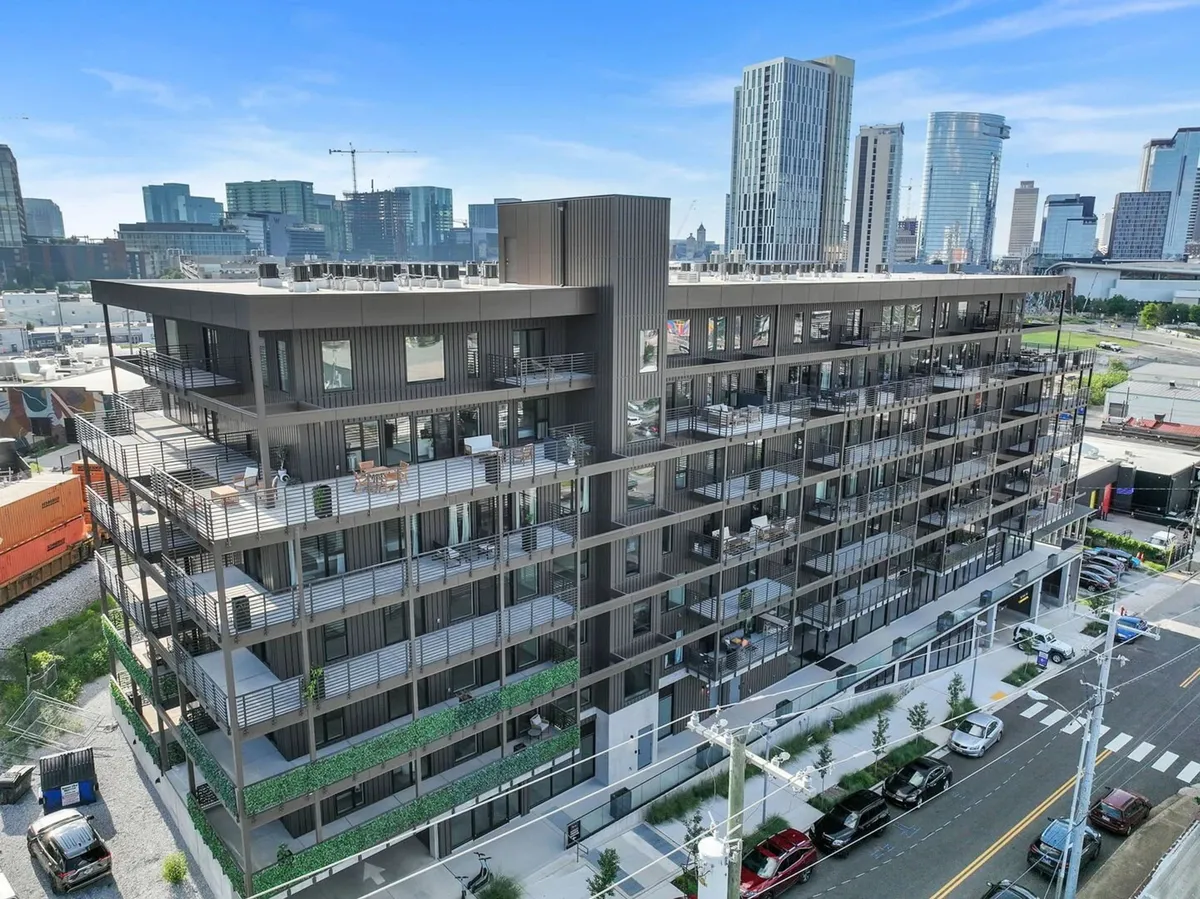 The exterior of the modern apartment building is captured, featuring balconies with comfortable outdoor furniture and a rooftop terrace ideal for gatherings. The lively and contemporary design invites guests to explore urban amenities or relax in communal outdoor spaces, with the vibrant neighborhood just beyond.