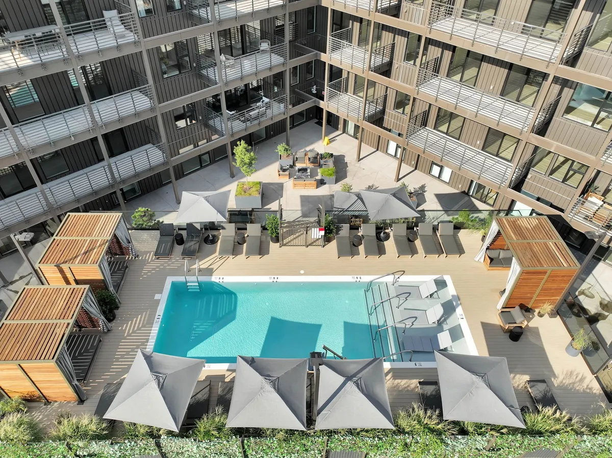 A vibrant exterior shot of the Hyve's sparkling swimming pool, surrounded by stylish lounge chairs, shaded canopies, and lush garden areas. The tranquil ambiance and inviting poolside space suggest leisurely relaxation, sunbathing, and opportunities for social gatherings or outdoor dining.