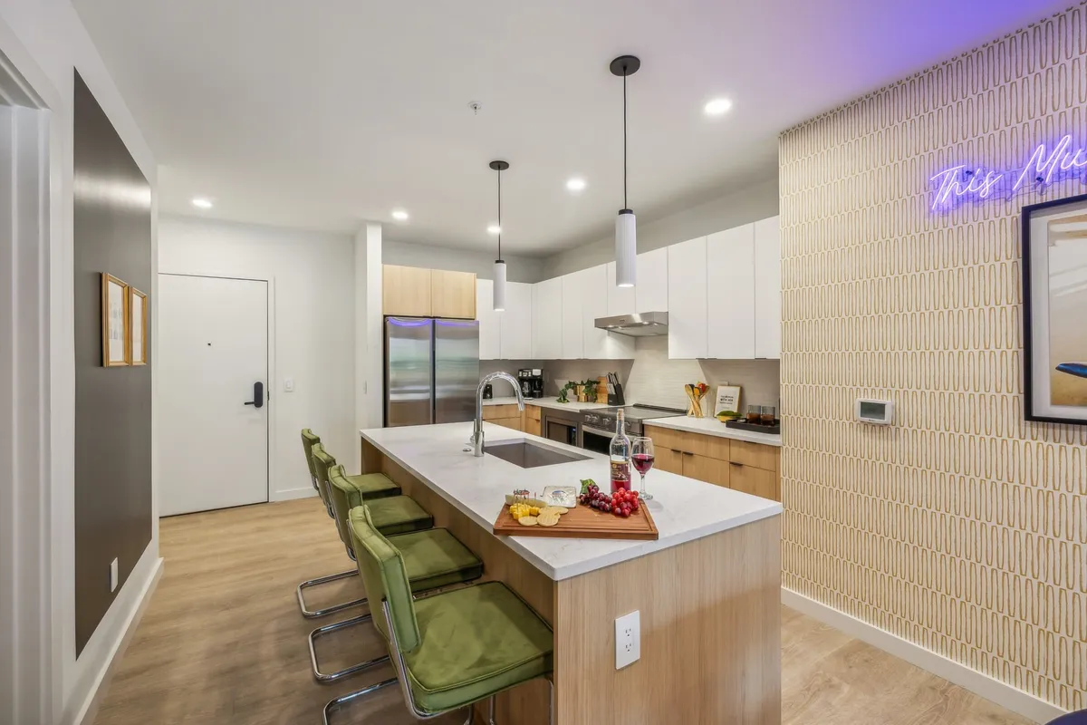 A sleek, modern kitchen with a prominent island, marble countertops, neon sign for a fun touch, and bar stools for casual dining or conversation. The wood cabinetry and open layout make it inviting for both cooking and entertaining.