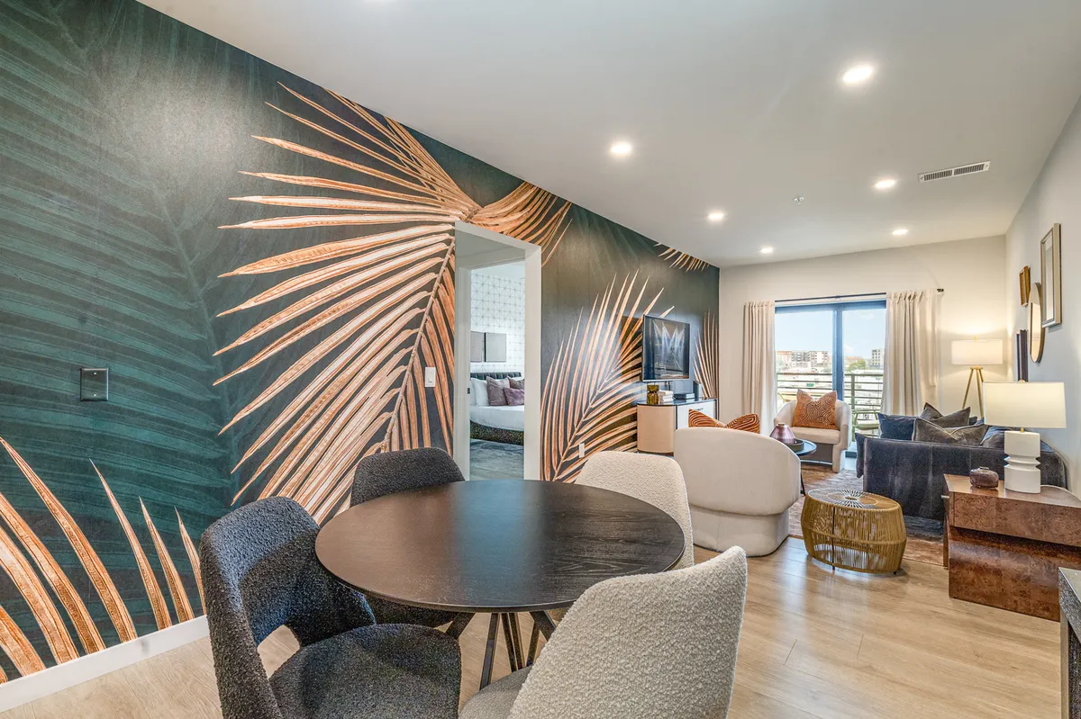 The image showcases a vibrant, contemporary living and dining area featuring a striking palm leaf mural, a spacious dining table perfect for group meals, a comfortable seating area for relaxation or social gatherings, and large windows that bathe the space in natural light. The scene exudes a fresh, inviting vibe designed for entertaining and unwinding.
