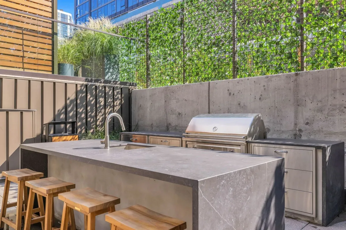 A modern outdoor kitchen area featuring a stainless steel grill, wooden stools, and a striking green wall, ideal for entertaining guests and hosting lively BBQs in the open air. The warmth of the wood and greenery provides a welcoming vibe.