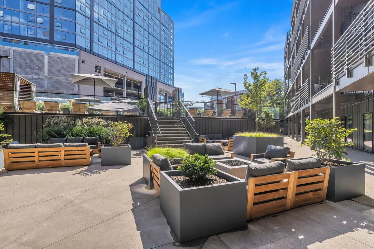 A rooftop lounge scene with comfortable seating areas, wooden planters, vibrant greenery, and a modern building facade. The staircase hints at upper-level amenities, while the inviting setup is perfect for gatherings or relaxing by the firepit under the city lights.