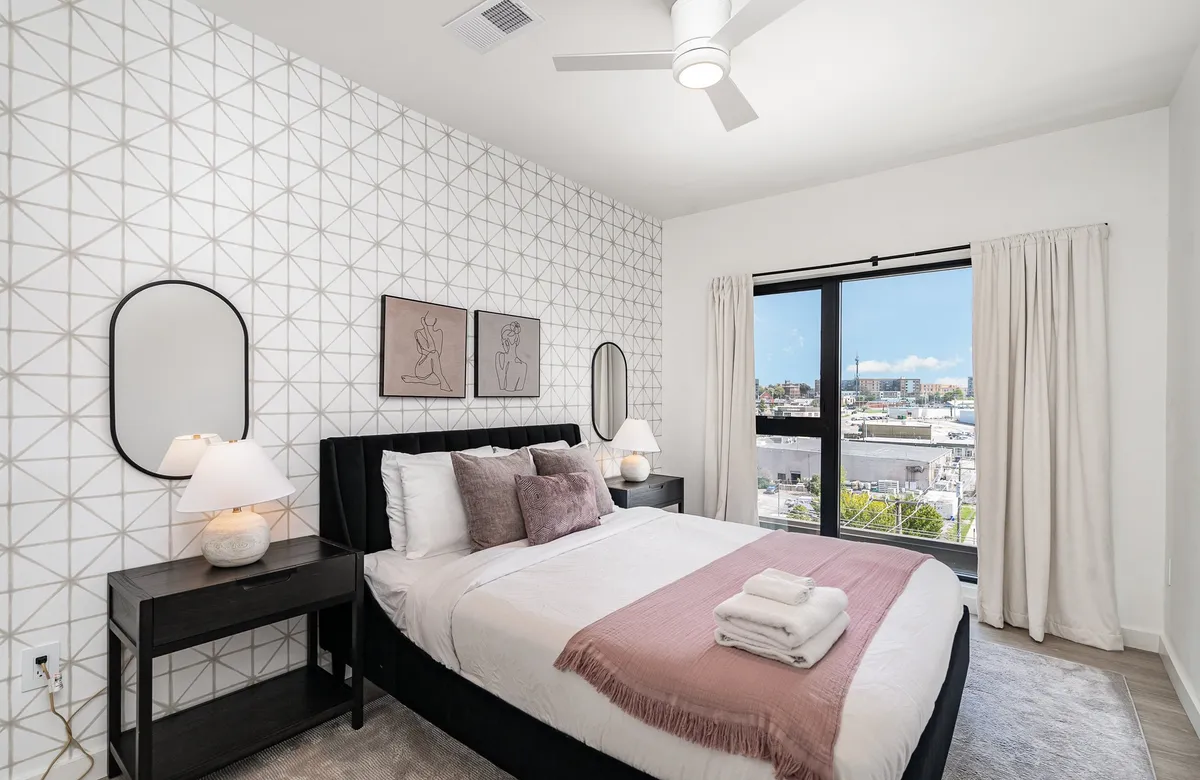 A serene bedroom with a large bed outfitted in plush pillows, a geometric accent wall adding modern flair, and a large window offering a peaceful city view. The décor is inviting, with soft natural light enhancing the tranquil atmosphere.