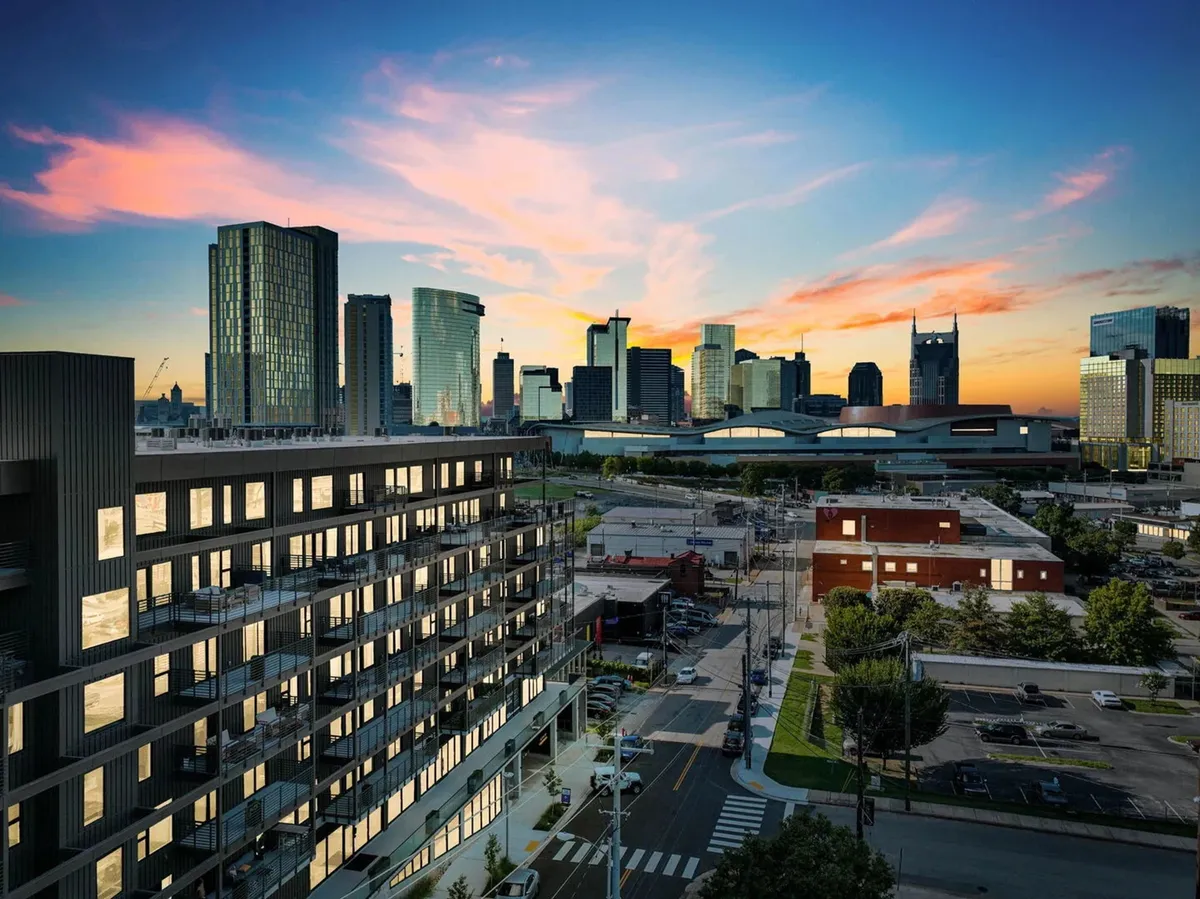 A modern apartment building is seen against the lively city skyline, illuminated by a colorful sunset. The vantage point highlights walkability to downtown attractions, with the cityscape and local venues visible in the background. This image captures the vibrant urban context and immediate access to Nashville’s entertainment and convention scene.