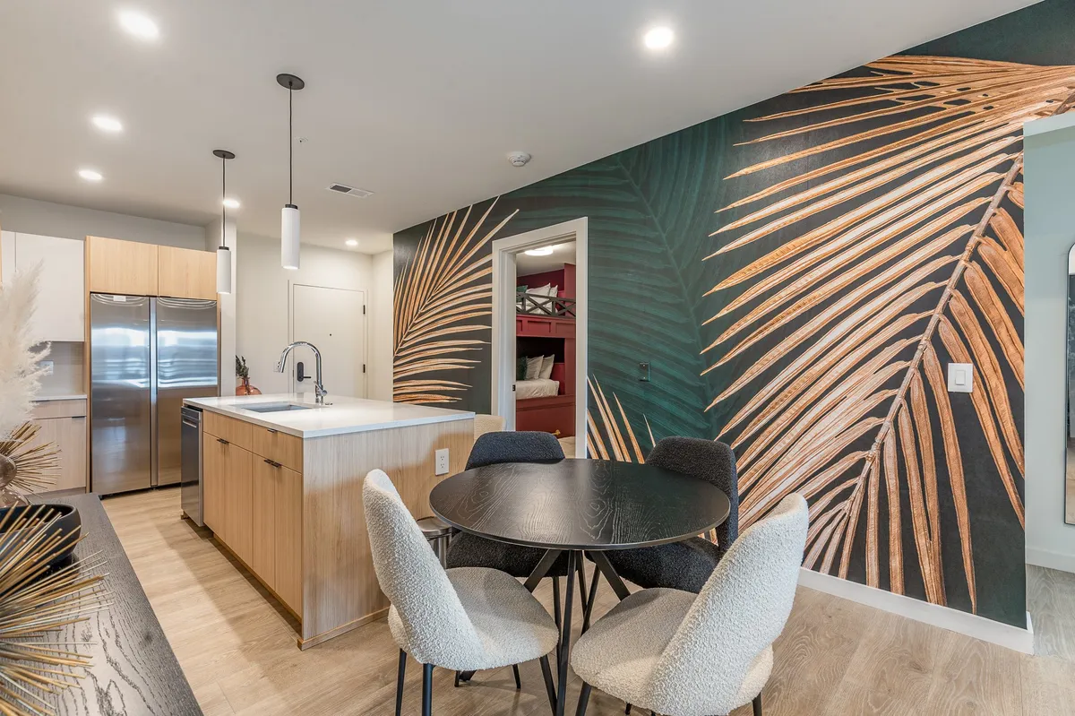 This image showcases a modern, open-concept space that artfully blends a kitchen island, dining table, and living area. The large palm leaf mural adds a lively, social vibe, and the furniture is stylish and contemporary. The kitchen is fully equipped with stainless steel appliances, and the overall layout invites group gatherings and social interactions.