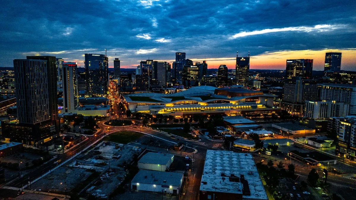 A panoramic view of the Nashville city skyline at sunset, with illuminated buildings and a warm glow highlighting the vibrancy of the neighborhood. The scene hints at the nearby entertainment, cultural landmarks, and family-friendly attractions that guests can easily walk to.