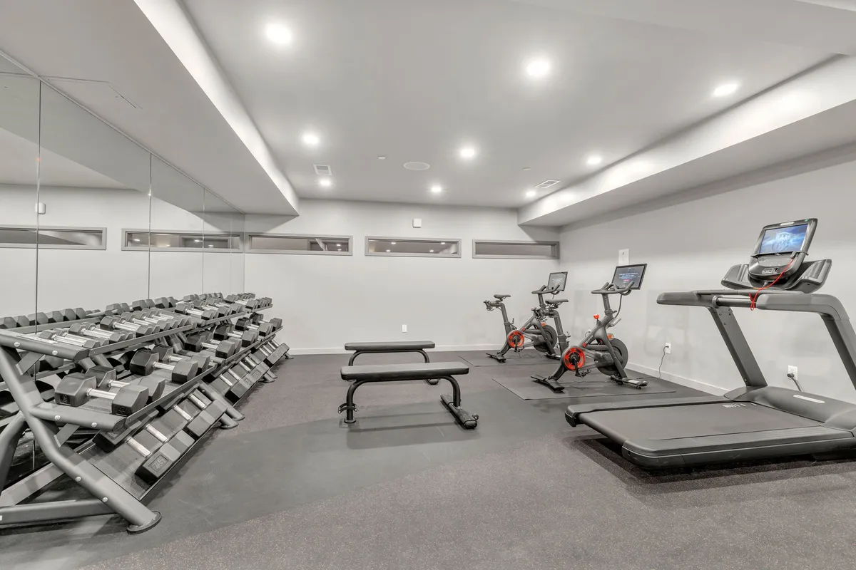 An interior view of the state-of-the-art fitness center, equipped with a dumbbell rack, treadmills, stationary bikes, and a weight bench. The motivational ambiance and modern equipment invite both solo workouts and group fitness sessions.